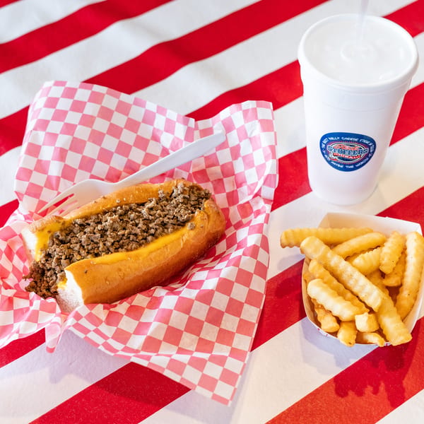 Cheesesteak in checkered paper, crinkle-cut fries, and a drink on a red and white striped table.