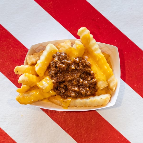 Chili cheese fries in a white paper tray, on a red and white striped background.