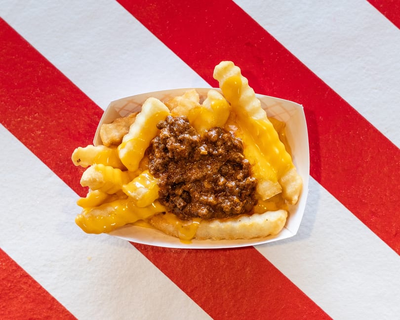 Chili cheese fries in a white paper tray, on a red and white striped background.
