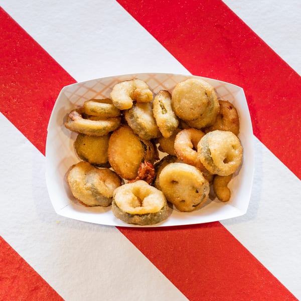 A white paper tray filled with golden-brown fried pickles, some round, some sliced, on a red and white striped surface.