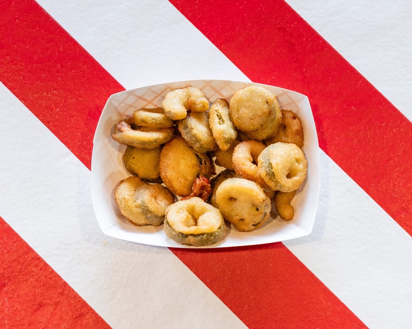 A white paper tray filled with golden-brown fried pickles, some round and some oval, on a red and white striped surface.