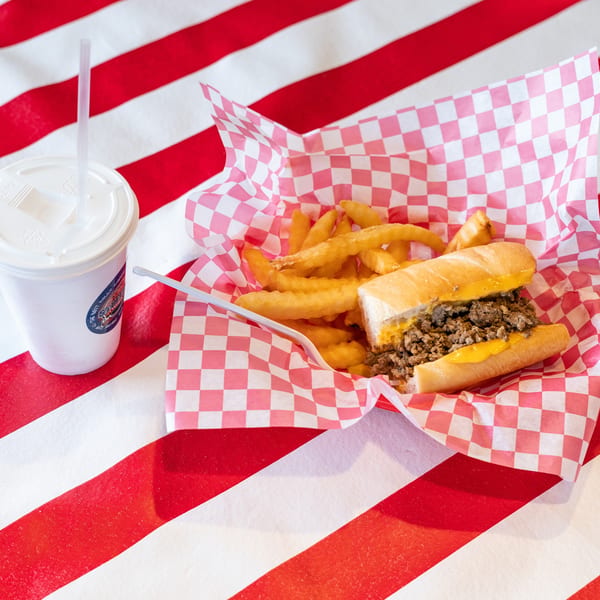 Cheesesteak sandwich and fries in a red checkered basket with a drink on a striped table.