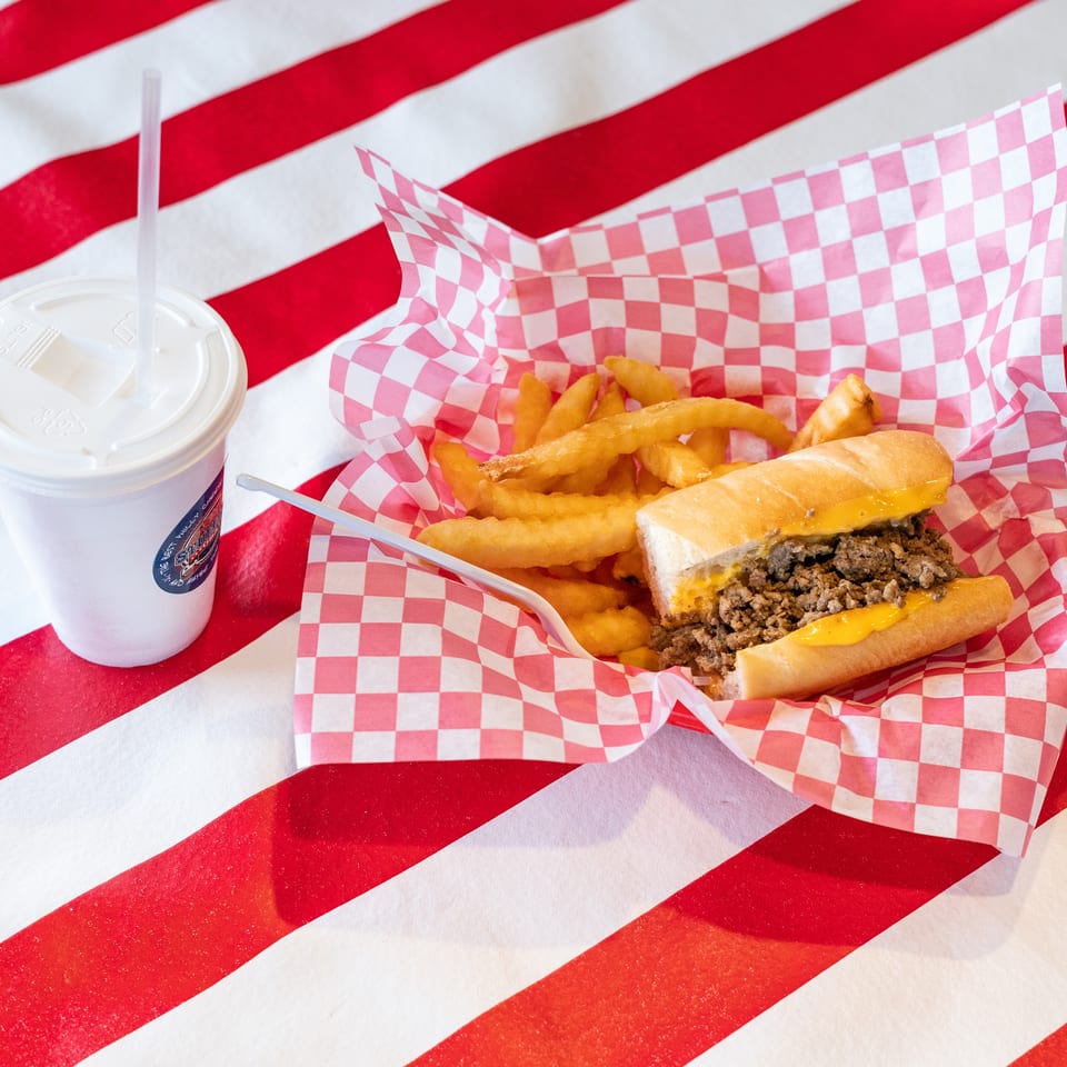Cheesesteak sandwich and fries in a red checkered basket with a drink on a striped table.