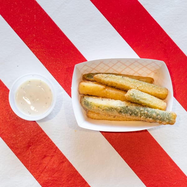 Fried pickle spears in a paper tray with a side of dipping sauce on a red and white striped surface.