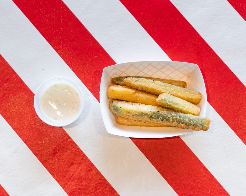 Fried pickle spears in a paper tray with a side of dipping sauce on a red and white striped surface.