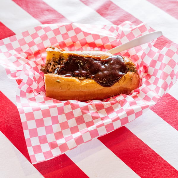 A chili dog with dark sauce and ground meat in a red and white checkered paper boat.