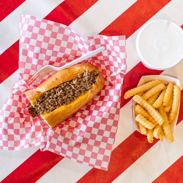 Cheesesteak sandwich with fries and a drink on a red and white striped background.