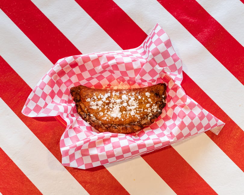 A golden-brown fried dessert empanada, dusted with powdered sugar, in a pink and white checkered paper.