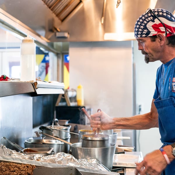 A chef in an American flag bandana stirs a steaming pot in a busy commercial kitchen.