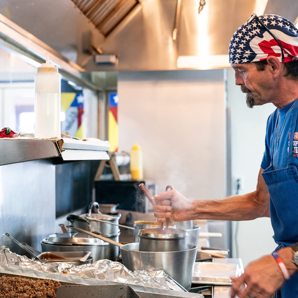 A chef in an American flag bandana stirs a steaming pot in a busy commercial kitchen.