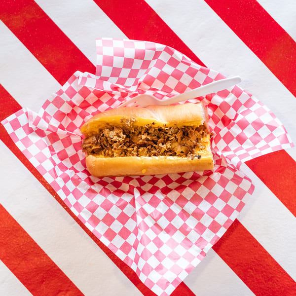 Cheesesteak sandwich with melted cheese and meat, served in checkered paper on a red and white striped background.