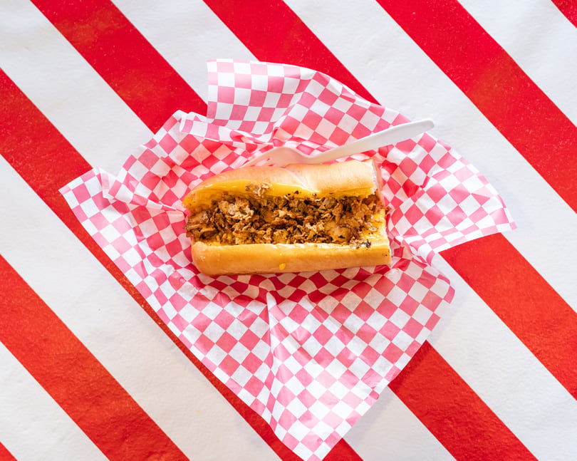 Cheesesteak sandwich with melted cheese and meat, served in checkered paper on a red and white striped background.