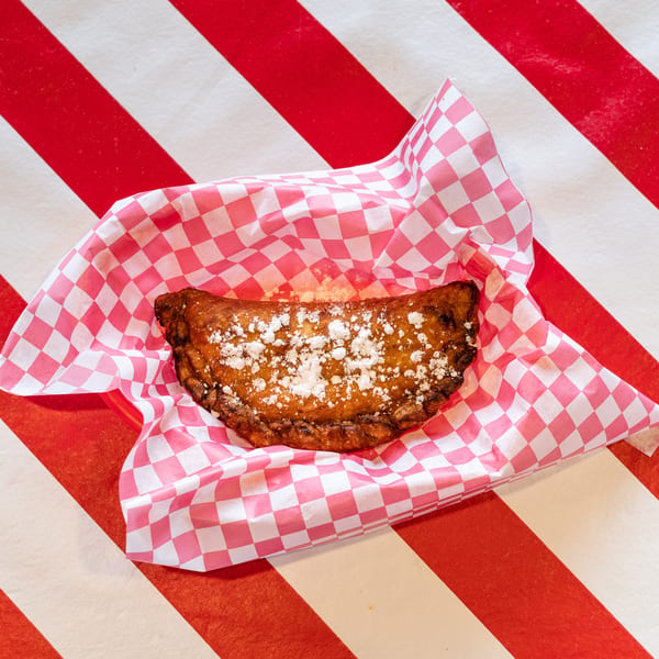 A golden-brown fried dessert empanada, dusted with powdered sugar, rests in a pink checkered paper.